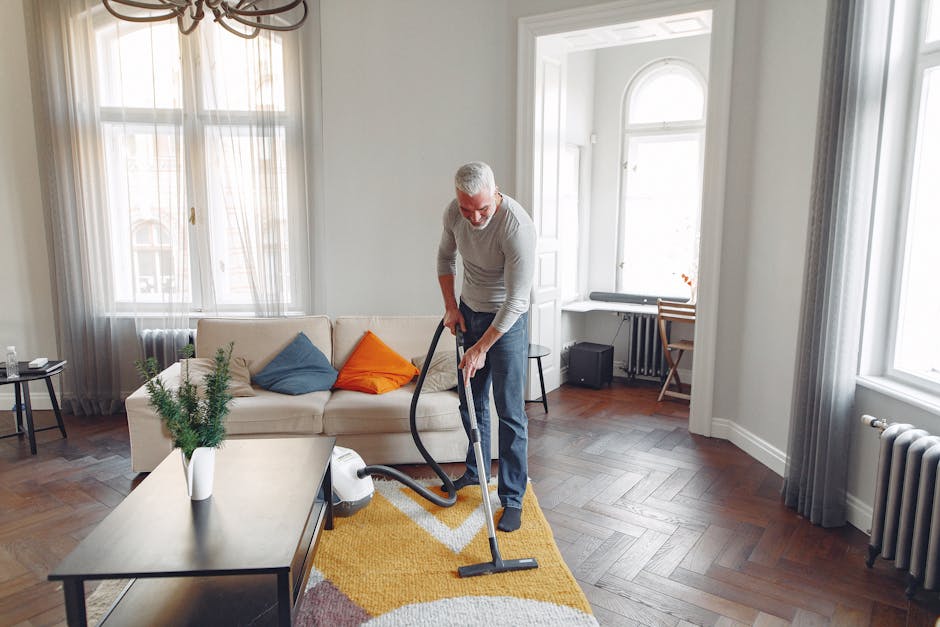 A man with grey hair and a beard using a vacuum cleaner to clean a yellow, white, and purple patterned rug in a bright, spacious living room. The room features a beige sofa with colorful cushions, a c