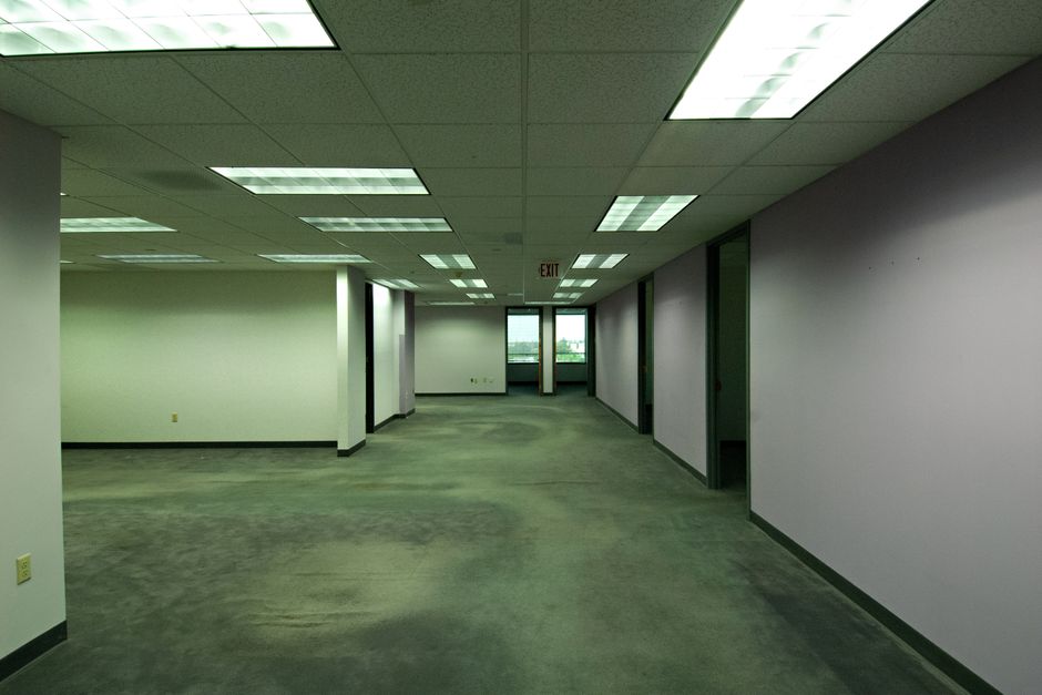 Close-up view of a commercial office space featuring a grey carpet floor with a textured surface, partially illuminated by natural light filtering through metallic horizontal blinds on large windows. 