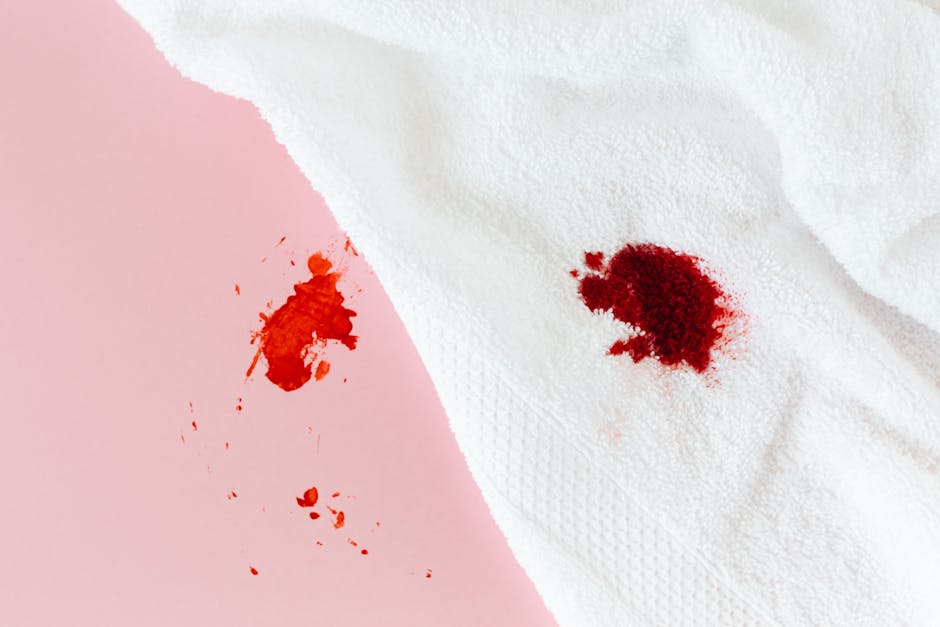 Close-up of a bedroom or living room surface with a white fabric garment, possibly a shirt, displaying a fresh red stain, likely blood. The fabric appears smooth and clean besides the stain, with natu