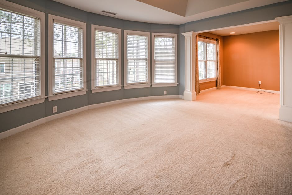 A person dressed in orange work overalls vacuuming a grey carpeted living room using a yellow commercial vacuum cleaner with a long black hose and a flat cleaning head. The room features a grey fabric