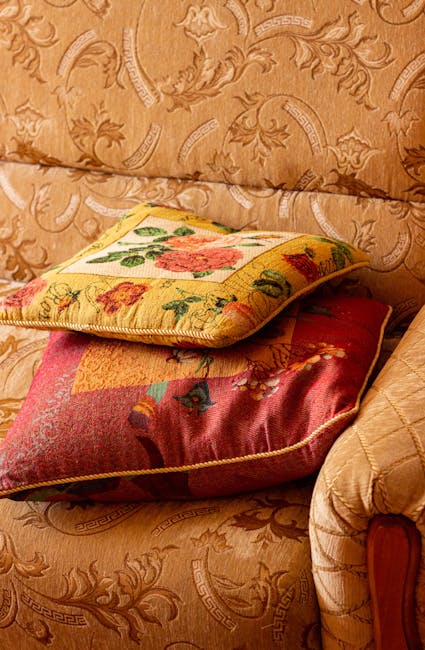 Close-up image of three decorative cushions placed on a vintage, ornately patterned, beige fabric sofa. The top cushion features a floral embroidery on a yellow background, with detailed stitching and vibrant flower motifs. The middle cushion has a quilted design with a multicolored, patchwork-like pattern, showcasing a mix of reds, pinks, and blues. The bottom cushion, partially visible, appears to have a plain, muted fabric with a slightly different texture. The sofa's fabric displays intricate, embossed scroll and leaf patterns, suggesting a period or vintage style. The soft lighting highlights the textures and the cleanliness of the cushions and fabric, indicating careful maintenance. This setting exemplifies the importance of surface cleaning and textile care in domestic environments, aligning with the focus of Surbiton KT6 upholstery cleaning guide for period homes, KINGSTON, as offered by Kingston Carpet Cleaning.