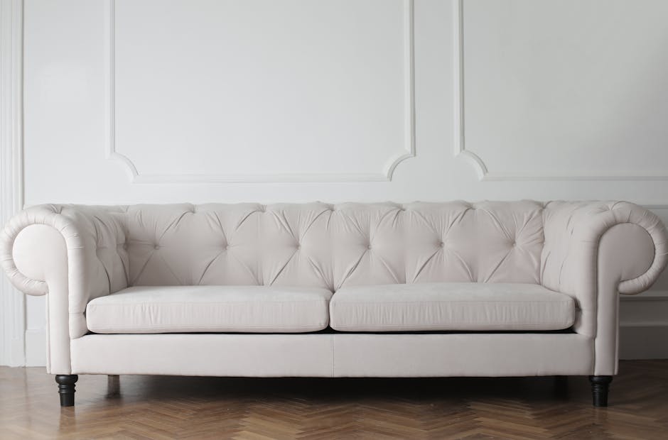A cream-colored, tufted fabric sofa with rolled arms sits against a white paneled wall in a well-lit room. The sofa's surface appears clean and smooth, with no visible dust or stains, indicating recent surface cleaning or maintenance. The floor is hardwood with a natural finish, and the lighting creates soft shadows, highlighting the sofa's upholstery and the elegant wall design. The overall scene suggests an environment that has been carefully cleaned and maintained, consistent with domestic surface cleaning and upholstery sanitisation services offered by Kingston Carpet Cleaning.