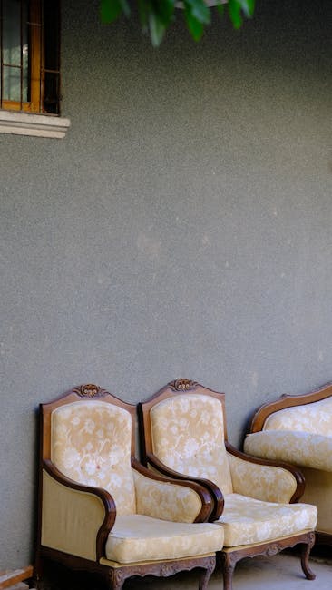 Close-up image of three decorative cushions placed on a vintage, ornately patterned, beige fabric sofa. The top cushion features a floral embroidery on a yellow background, with detailed stitching and vibrant flower motifs. The middle cushion has a quilted design with a multicolored, patchwork-like pattern, showcasing a mix of reds, pinks, and blues. The bottom cushion, partially visible, appears to have a plain, muted fabric with a slightly different texture. The sofa's fabric displays intricate, embossed scroll and leaf patterns, suggesting a period or vintage style. The soft lighting highlights the textures and the cleanliness of the cushions and fabric, indicating careful maintenance. This setting exemplifies the importance of surface cleaning and textile care in domestic environments, aligning with the focus of Surbiton KT6 upholstery cleaning guide for period homes, KINGSTON, as offered by Kingston Carpet Cleaning.