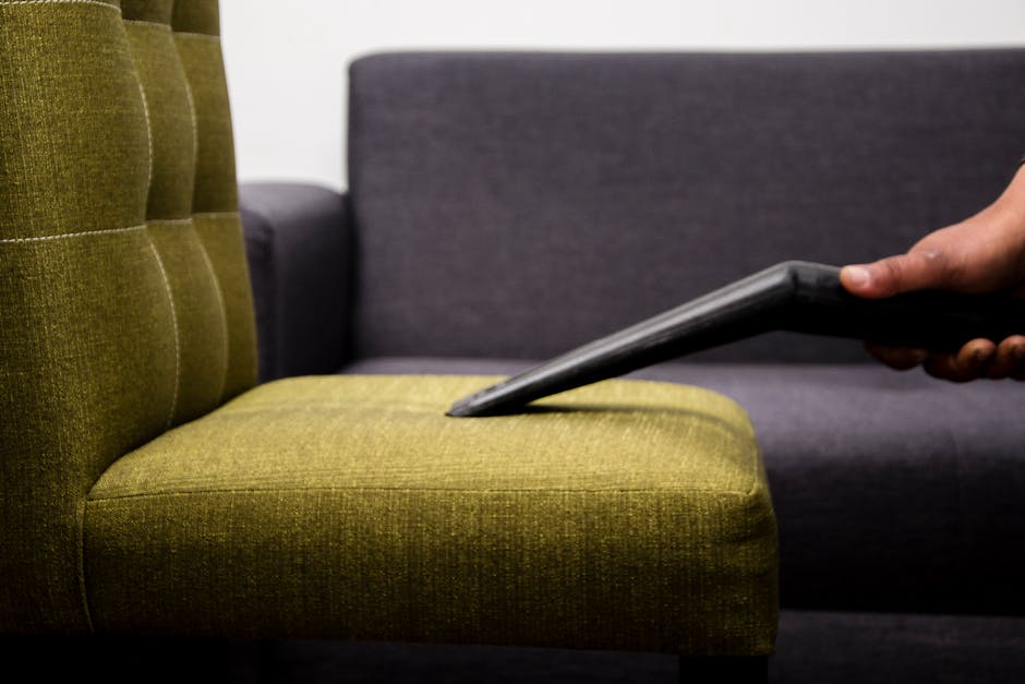 Close-up of a decorative cushion with a leaf pattern in shades of black, gray, and blue, placed on a gray fabric sofa in a living room. The sofa has a textured surface and is positioned against a bric