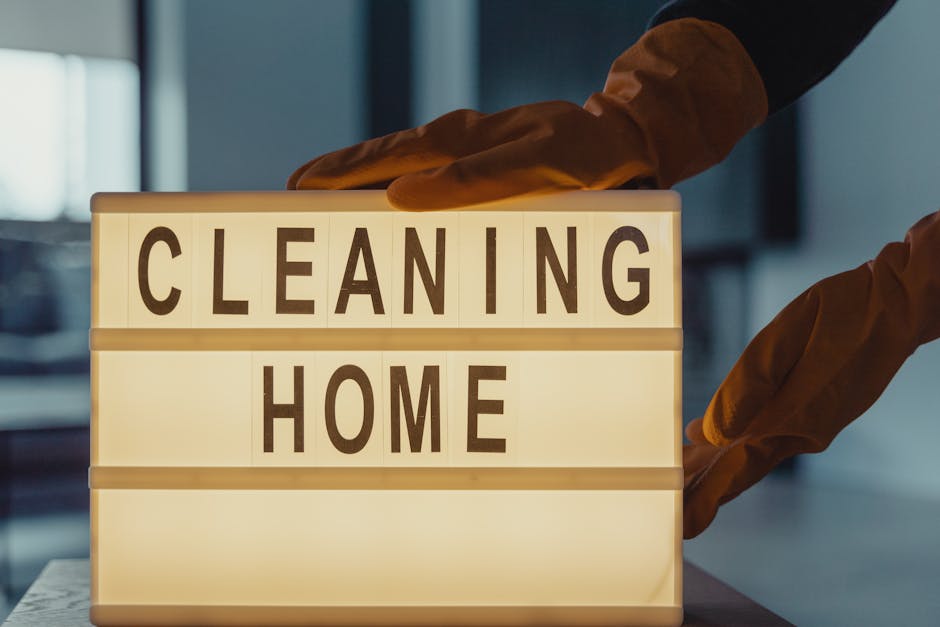 A person wearing brown cleaning gloves is placing a lit-up sign with the words 'CLEANING HOME' in black capital letters on a flat surface. The background appears to be an indoor space with natural light coming from a window, and the surface of the table looks clean and smooth. The lighting highlights the sign prominently, emphasizing the theme of domestic cleaning services offered by Kingston Carpet Cleaning for end of tenancy cleaning in Kingston town centre.