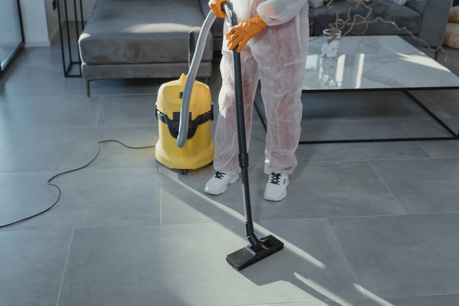 A person dressed in white protective coveralls, orange gloves, and white sneakers is engaged in surface cleaning on a large, grey tiled floor in a modern living room. They are using a vacuum cleaner with a grey hose, connected to a yellow upright extractor machine placed on the floor nearby. The room features a grey fabric sofa, a glass-topped coffee table, and a metal side table, with ample natural light streaming in. The tiles appear clean and well-maintained, with a smooth, shiny finish indicative of thorough deep cleaning and sanitisation, typical of professional end-of-tenancy cleaning services provided by Kingston Carpet Cleaning. The overall scene highlights the meticulous cleaning process for preparing a property for new occupancy, emphasizing hygiene and surface maintenance.